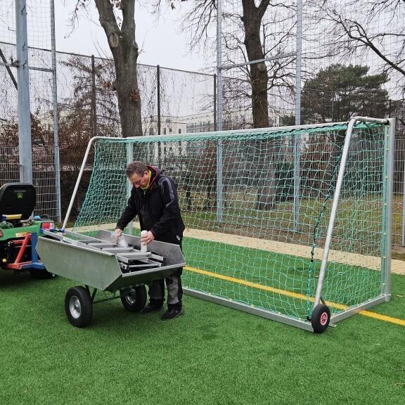 Ein Mann arbeitet an einer mobilen Tor-Ausr&uuml;stung neben einem kleinen gr&uuml;nen Spielzeugtraktor auf einem Sportplatz.