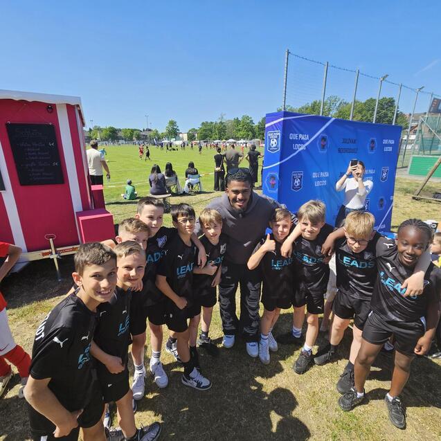 Gruppe von Kindern in Sportbekleidung posiert l&auml;chelnd mit einem Erwachsenen vor einem Fu&szlig;ballspiel im Freien.