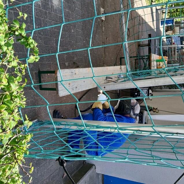 Arbeiter in Schutzkleidung, der an einem Ger&uuml;st arbeitet, mit einem Holzbrett und gr&uuml;nen Netzen im Vordergrund.