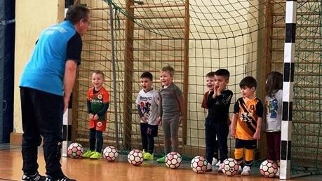 Ein Trainer spricht mit einer Gruppe von Kindern, die im Tor stehen, umringt von Fußbällen in einer Sporthalle.