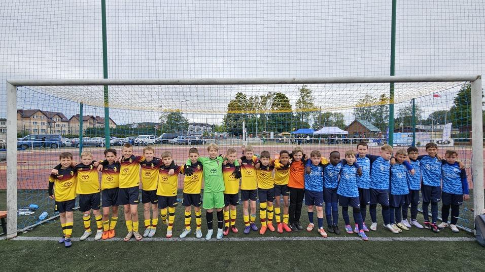 Gruppenshot von zwei Jugendfußballmannschaften in Trikots, stehend vor einem Tor auf einem Kunstrasenplatz.