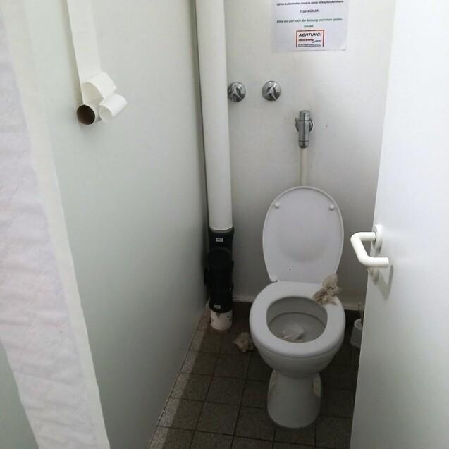 Toilettenraum mit einem Klo, grauen Fliesen und einem Hinweisblatt an der Wand. Sichtbare Abfallstoffe auf dem Boden.
