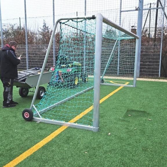 Mann mit Handy vor einem Fußballtor auf einem Kunstrasenplatz, im Hintergrund ein Wagen mit Sportgeräten.