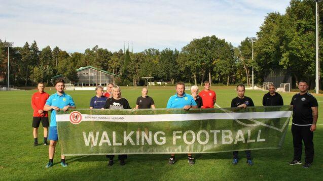 Gruppe von elf Personen auf einem Fußballplatz, die ein Banner mit der Aufschrift "WALKING FOOTBALL" halten.