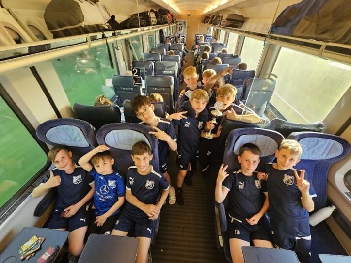 Gruppe von Jungen in Sportkleidung sitzt im Zug, einige zeigen V-Zeichen, einer hält einen Pokal.