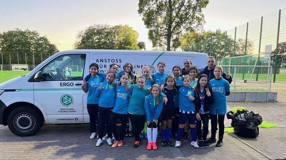 Gruppe von Kindern in Sportkleidung steht lächelnd vor einem Fußballbus auf einem Sportplatz.