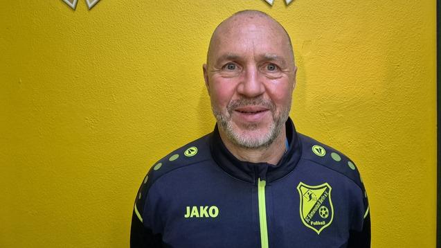 Porträt eines lächelnden Mannes in Sportjacke vor gelber Wand mit Fußball-Trophäen im Hintergrund.