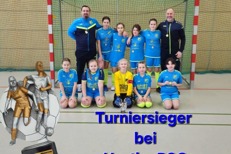 M&auml;dchen-Fu&szlig;ballmannschaft in blauen Trikots mit Trainern vor einem Tor, daneben eine Pokal-Statue und der Text &bdquo;Turniersieger bei Hertha BSC&ldquo;.