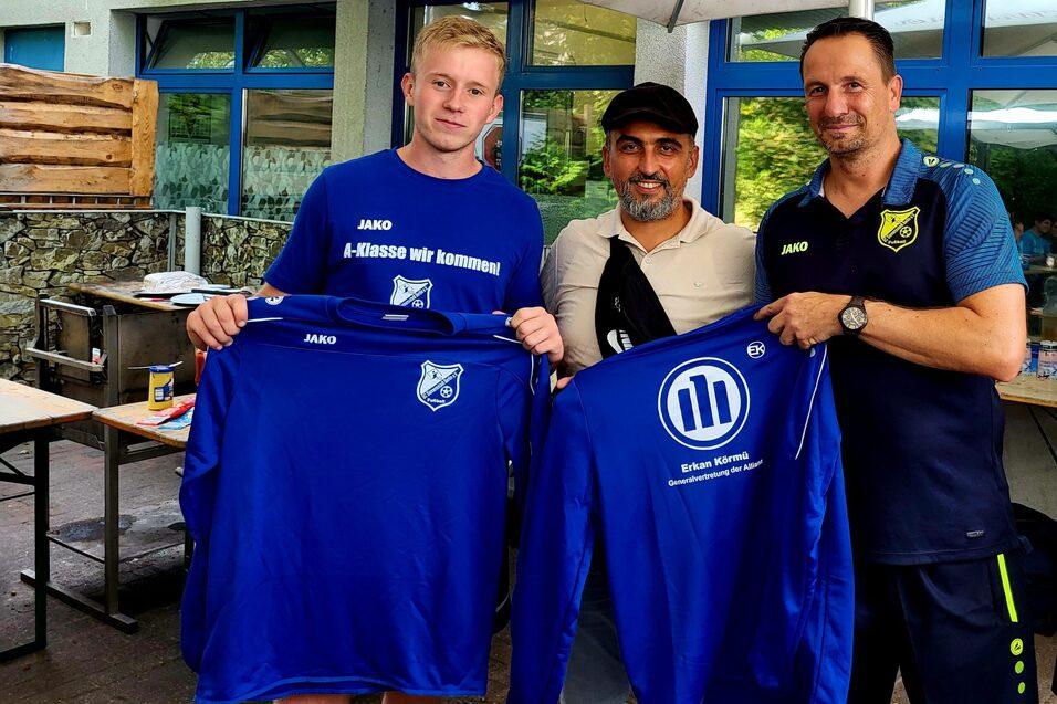 Drei M&auml;nner halten Fu&szlig;balltrikots in blauer Farbe, w&auml;hrend sie in einem Gartenbereich stehen.