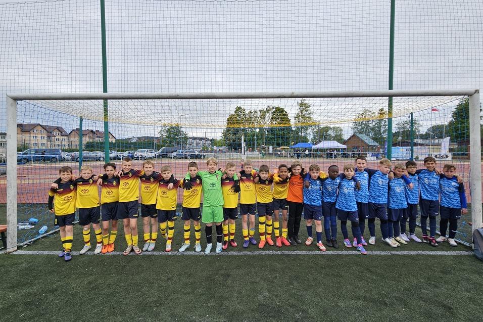 Gruppenshot von zwei Jugendfu&szlig;ballmannschaften in Trikots, stehend vor einem Tor auf einem Kunstrasenplatz.