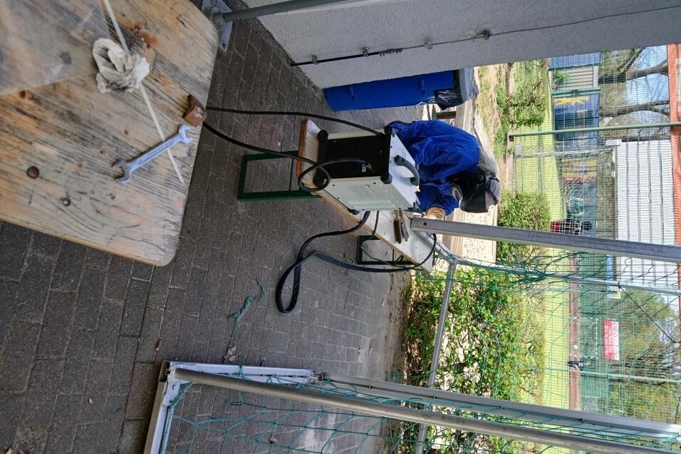 Arbeiter in blauer Schutzkleidung montiert Ger&auml;te an einem Tisch im Freien, umgeben von einer Sportanlage.