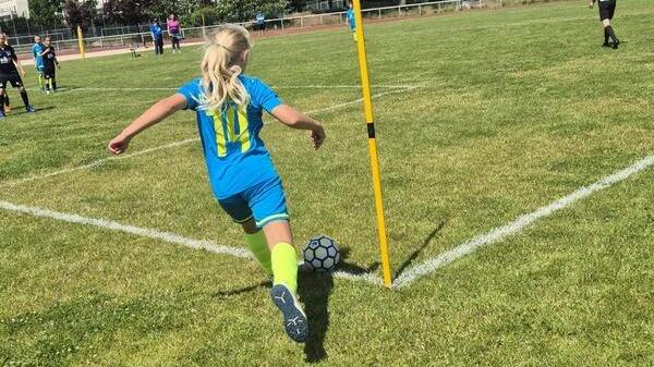 Ein Mädchen in blau-grünem Fußballtrikot führt einen Eckball auf einem Rasenplatz aus, umgeben von Spielern.