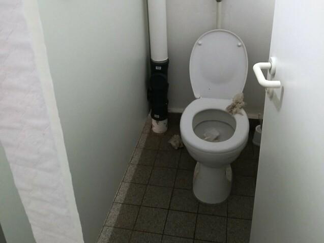 Toilettenraum mit einem Klo, grauen Fliesen und einem Hinweisblatt an der Wand. Sichtbare Abfallstoffe auf dem Boden.