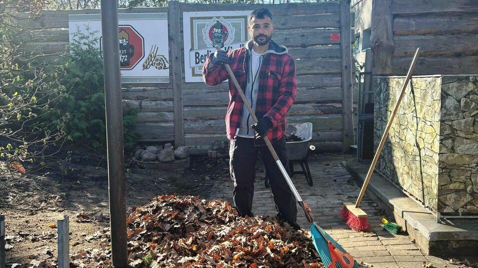Mann mit Rechen steht neben einem großen Haufen Laub in einem Garten, umgeben von Bäumen und Holzlattenzaun.