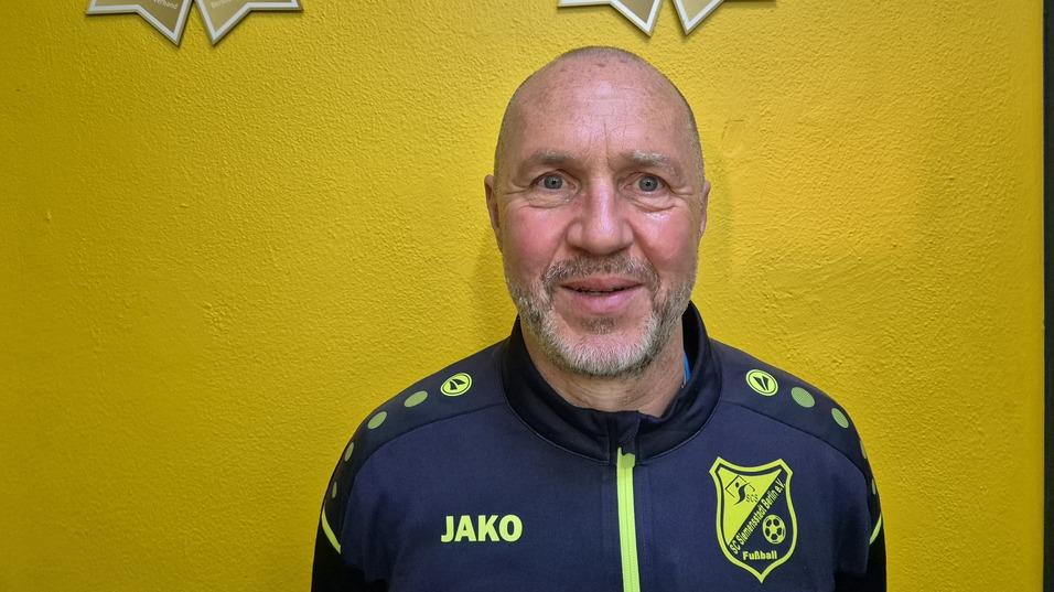 Porträt eines lächelnden Mannes in Sportjacke vor gelber Wand mit Fußball-Trophäen im Hintergrund.