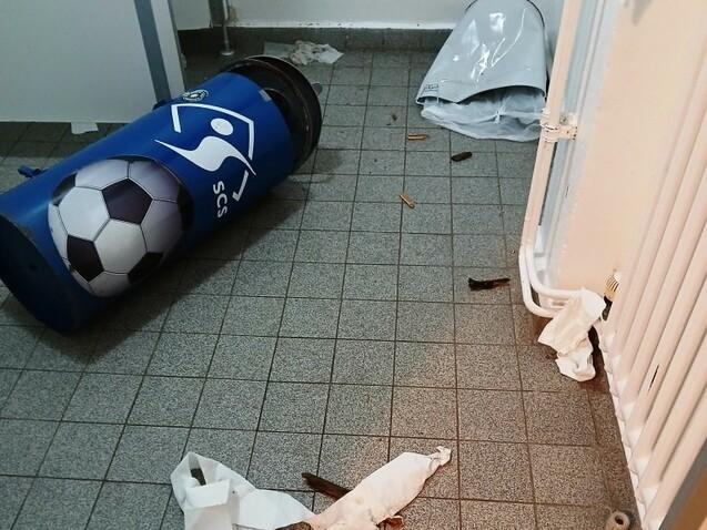Müll und Papier liegen verstreut auf einem Fliesenboden, ein umgefallenes Müllfass mit Fußballmotiv ist sichtbar.