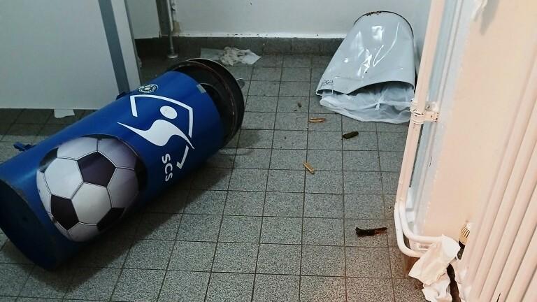 Müll und Papier liegen verstreut auf einem Fliesenboden, ein umgefallenes Müllfass mit Fußballmotiv ist sichtbar.
