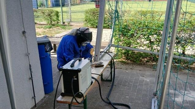 Arbeiter in blauer Schutzkleidung montiert Geräte an einem Tisch im Freien, umgeben von einer Sportanlage.