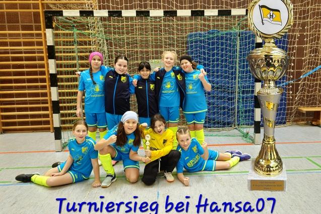 M&auml;dchenfu&szlig;ballmannschaft posiert vor einem Pokal in einer Sporthalle, alle tragen blaue Trikots mit gelben Akzenten.