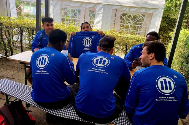 Gruppe von M&auml;nnern in blauen Shirts mit "Erkan K&ouml;rm&uuml;" am Tisch, w&auml;hrend einer ein Shirt pr&auml;sentiert.