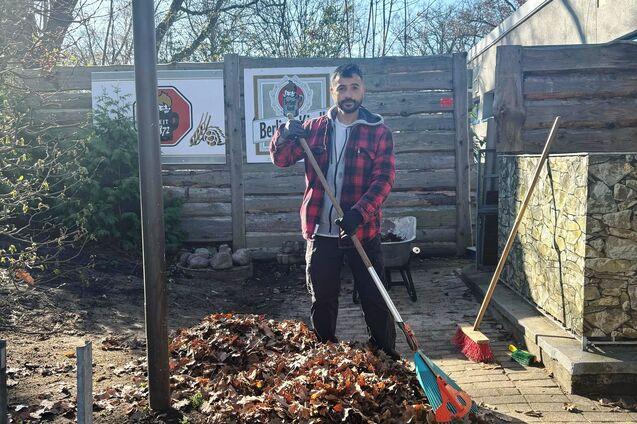 Mann mit Rechen steht neben einem gro&szlig;en Haufen Laub in einem Garten, umgeben von B&auml;umen und Holzlattenzaun.