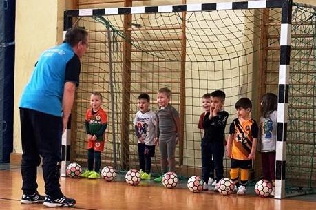 Ein Trainer spricht mit einer Gruppe von Kindern, die im Tor stehen, umringt von Fu&szlig;b&auml;llen in einer Sporthalle.