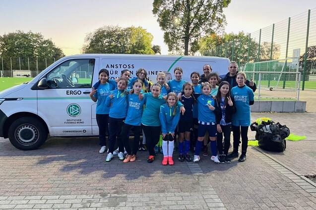 Gruppe von Kindern in Sportkleidung steht l&auml;chelnd vor einem Fu&szlig;ballbus auf einem Sportplatz.