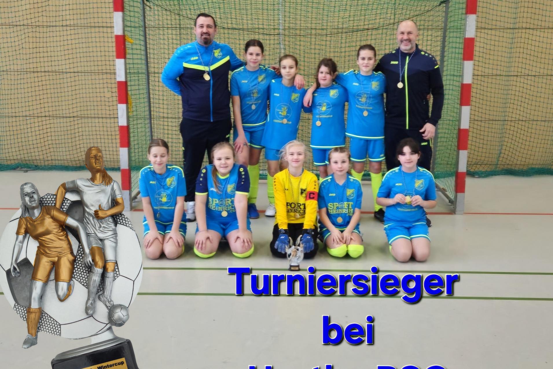 M&auml;dchen-Fu&szlig;ballmannschaft in blauen Trikots mit Trainern vor einem Tor, daneben eine Pokal-Statue und der Text &bdquo;Turniersieger bei Hertha BSC&ldquo;.