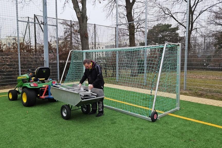 Ein Mann arbeitet an einer mobilen Tor-Ausr&uuml;stung neben einem kleinen gr&uuml;nen Spielzeugtraktor auf einem Sportplatz.