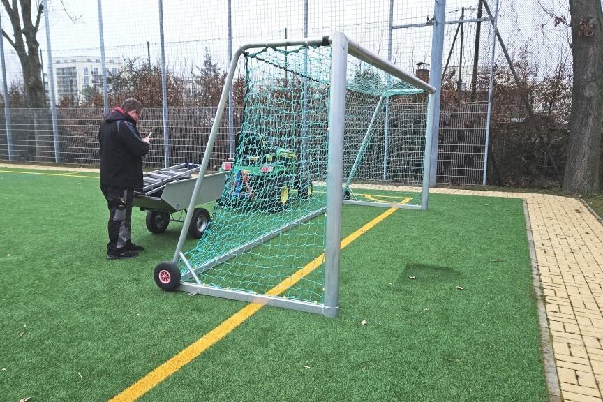 Mann mit Handy vor einem Fu&szlig;balltor auf einem Kunstrasenplatz, im Hintergrund ein Wagen mit Sportger&auml;ten.