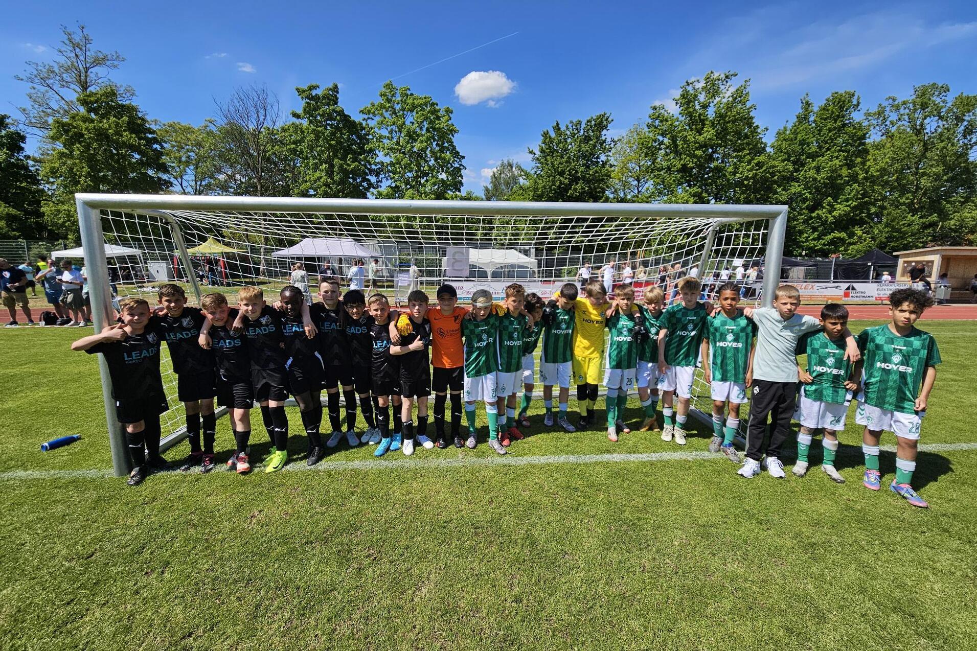 Zwei Fu&szlig;ballmannschaften stehen nebeneinander vor einem Tor auf einem gr&uuml;nen Spielfeld, umgeben von B&auml;umen und Zuschauern.