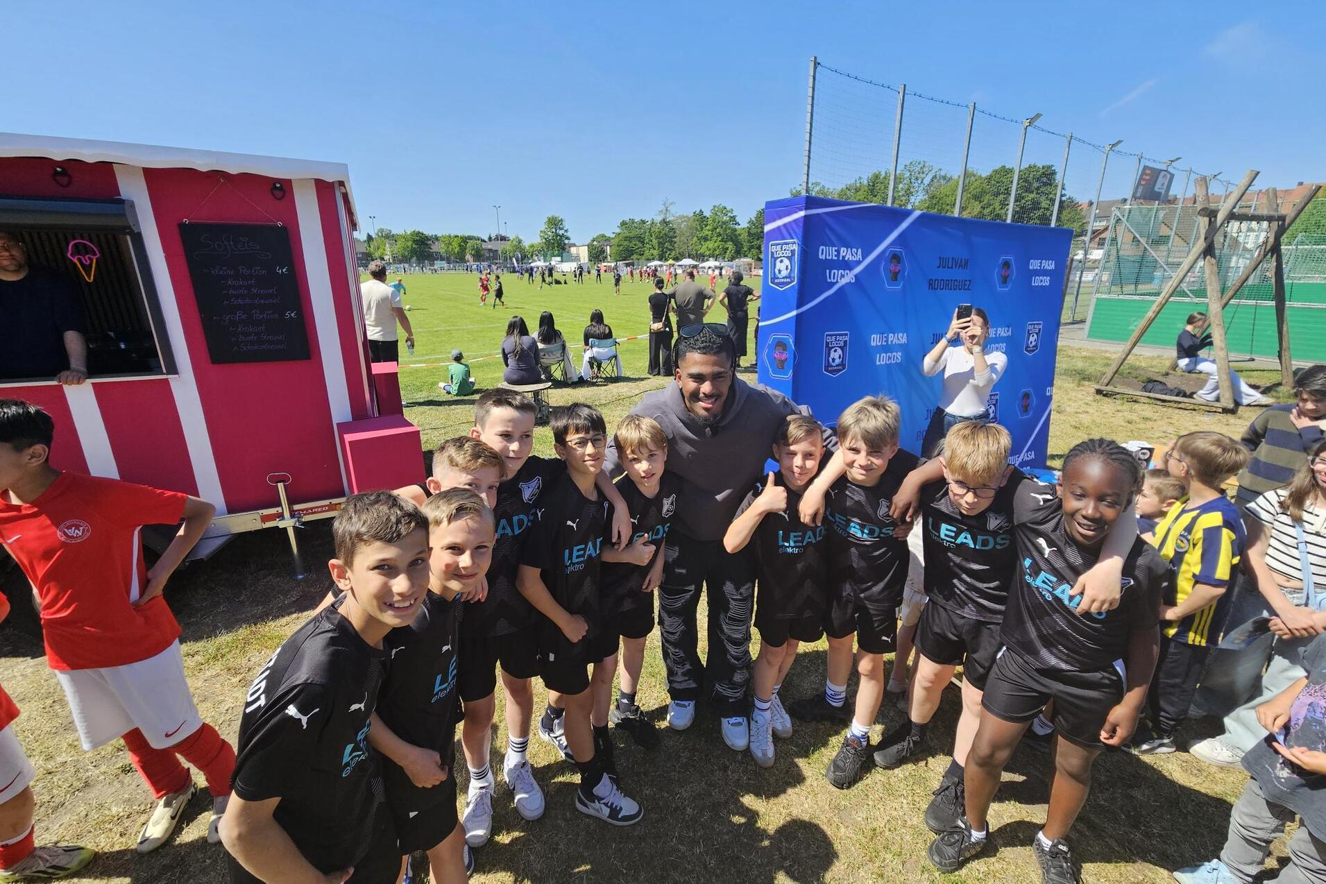 Gruppe von Kindern in Sportbekleidung posiert l&auml;chelnd mit einem Erwachsenen vor einem Fu&szlig;ballspiel im Freien.