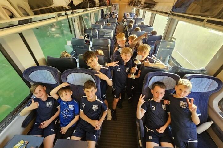 Gruppe von Jungen in Sportkleidung sitzt im Zug, einige zeigen V-Zeichen, einer h&auml;lt einen Pokal.