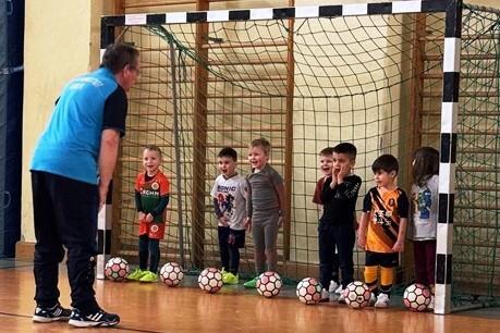 Ein Trainer spricht mit einer Gruppe von Kindern, die im Tor stehen, umringt von Fu&szlig;b&auml;llen in einer Sporthalle.