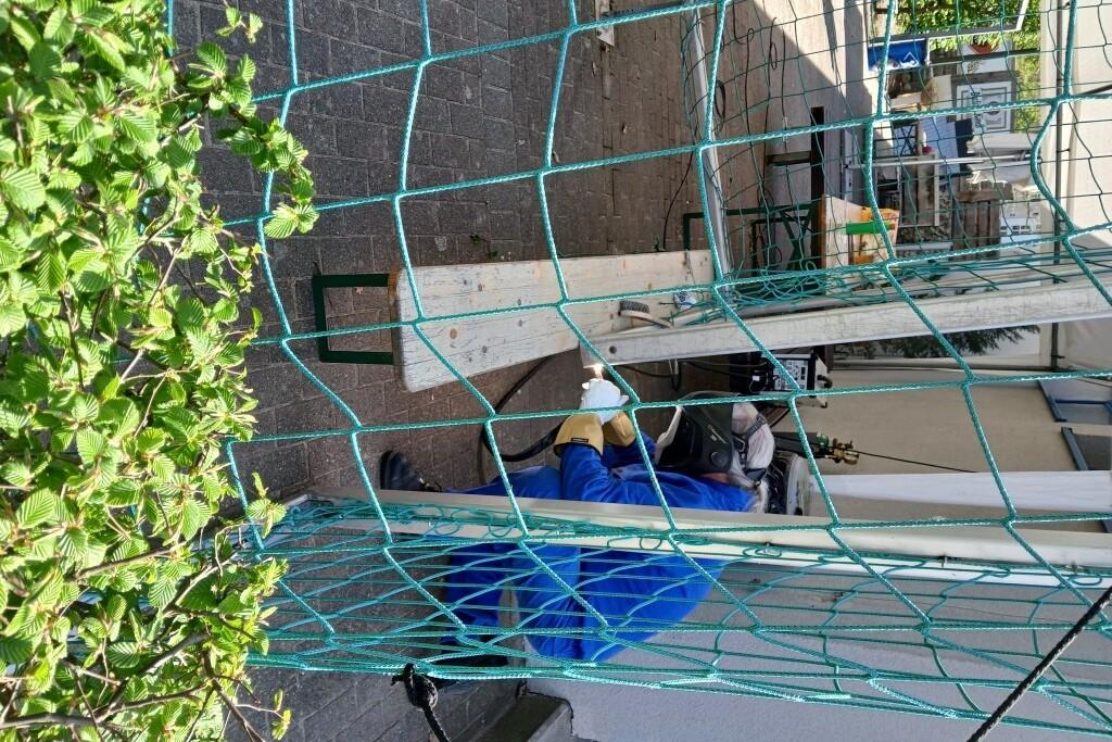 Arbeiter in Schutzkleidung, der an einem Ger&uuml;st arbeitet, mit einem Holzbrett und gr&uuml;nen Netzen im Vordergrund.