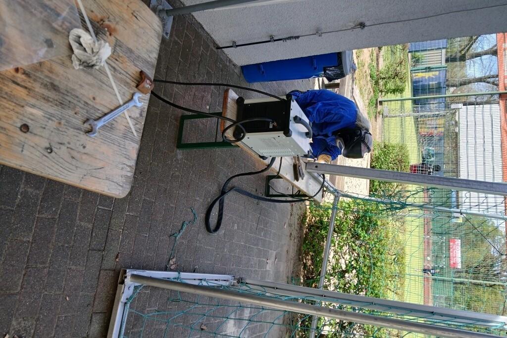 Arbeiter in blauer Schutzkleidung montiert Ger&auml;te an einem Tisch im Freien, umgeben von einer Sportanlage.