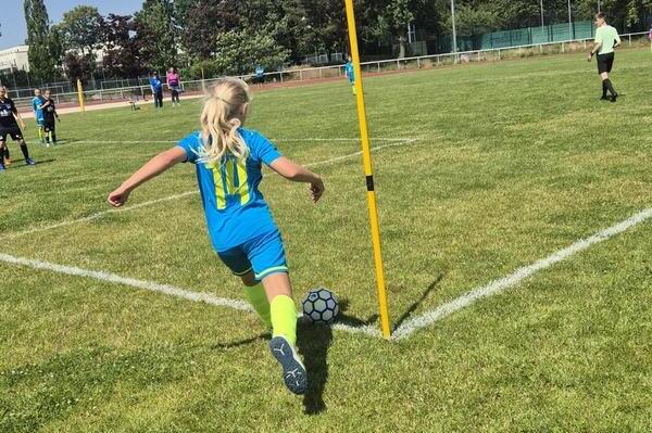 Ein Mädchen in blau-grünem Fußballtrikot führt einen Eckball auf einem Rasenplatz aus, umgeben von Spielern.