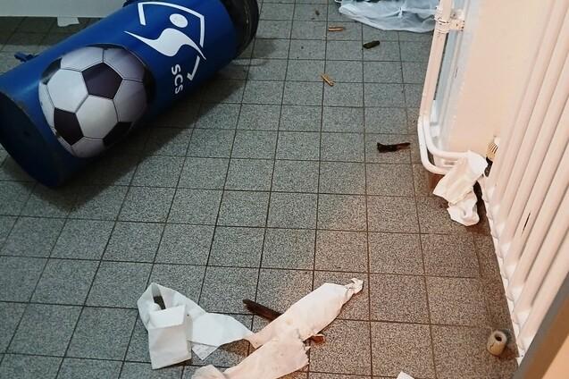 Müll und Papier liegen verstreut auf einem Fliesenboden, ein umgefallenes Müllfass mit Fußballmotiv ist sichtbar.