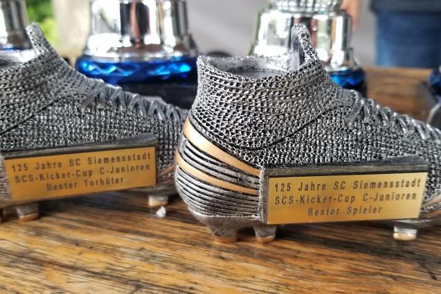 Zwei Fußballschuhe als Trophäen mit Namensplaketten „Bester Torhüter“ und „Bester Spieler“ auf einer Holzoberfläche.