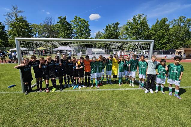 Zwei Fußballmannschaften stehen nebeneinander vor einem Tor auf einem grünen Spielfeld, umgeben von Bäumen und Zuschauern.