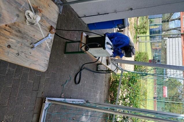 Arbeiter in blauer Schutzkleidung montiert Geräte an einem Tisch im Freien, umgeben von einer Sportanlage.