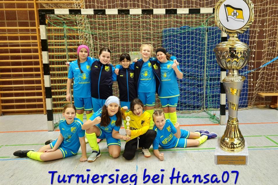 M&auml;dchenfu&szlig;ballmannschaft posiert vor einem Pokal in einer Sporthalle, alle tragen blaue Trikots mit gelben Akzenten.