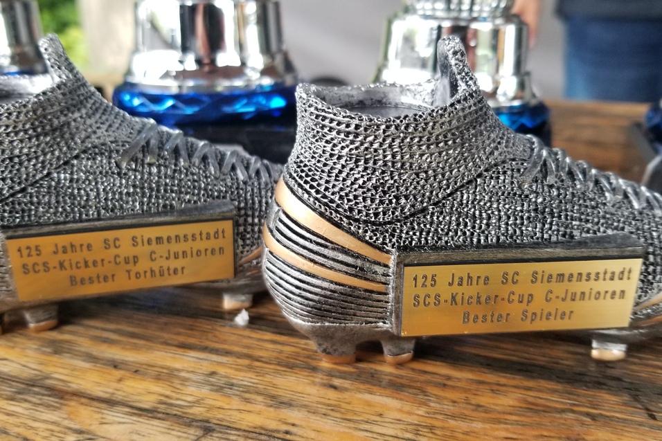 Zwei Fu&szlig;ballschuhe als Troph&auml;en mit Namensplaketten &bdquo;Bester Torh&uuml;ter&ldquo; und &bdquo;Bester Spieler&ldquo; auf einer Holzoberfl&auml;che.