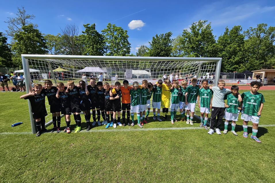 Zwei Fu&szlig;ballmannschaften stehen nebeneinander vor einem Tor auf einem gr&uuml;nen Spielfeld, umgeben von B&auml;umen und Zuschauern.