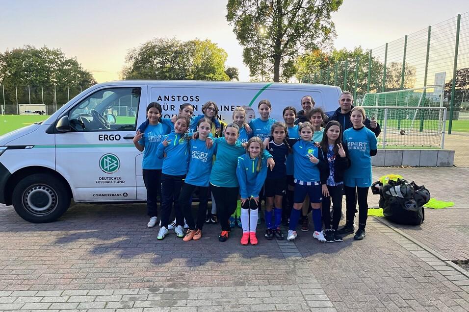 Gruppe von Kindern in Sportkleidung steht l&auml;chelnd vor einem Fu&szlig;ballbus auf einem Sportplatz.