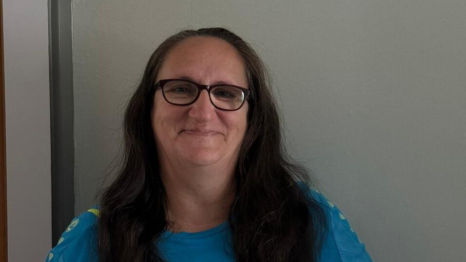 Eine lächelnde Frau mit langen, lockigen Haaren trägt ein blaues T-Shirt und sitzt vor einer grauen Wand.