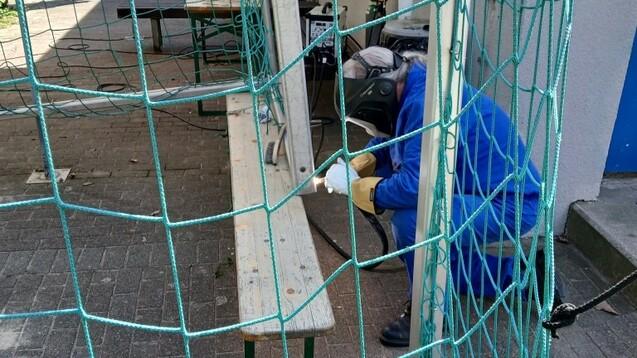 Arbeiter in Schutzkleidung, der an einem Gerüst arbeitet, mit einem Holzbrett und grünen Netzen im Vordergrund.