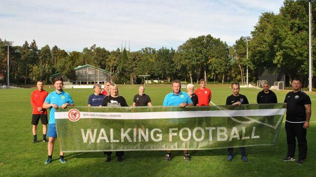 Gruppe von elf Personen auf einem Fu&szlig;ballplatz, die ein Banner mit der Aufschrift "WALKING FOOTBALL" halten.