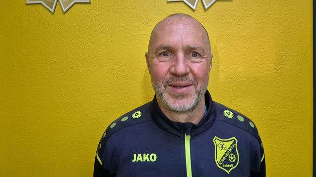 Porträt eines lächelnden Mannes in Sportjacke vor gelber Wand mit Fußball-Trophäen im Hintergrund.