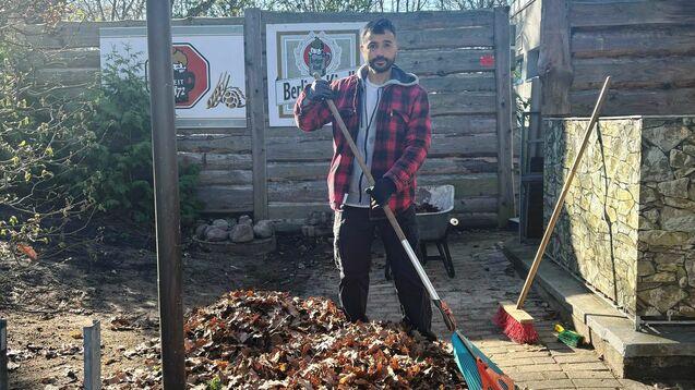 Mann mit Rechen steht neben einem großen Haufen Laub in einem Garten, umgeben von Bäumen und Holzlattenzaun.